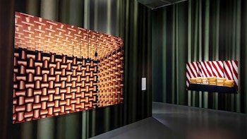 Dos obras de arte en una sala de exposición. Una con patrón cuadriculado marrón. La otra con rayas rojas y blancas, y pilas de documentos amarillos sobre mesa