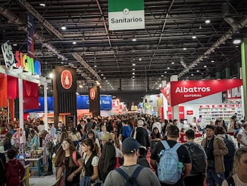 Interior de una convención con gran afluencia de gente. Se observan stands de editoriales como Penguin y Albatros, y señalización de 'Sanitarios'