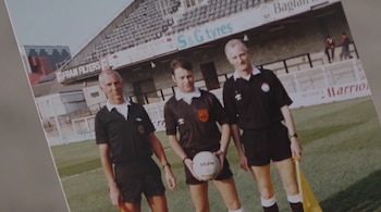 Fotografía de tres hombres vestidos de árbitros de fútbol. Uno sostiene un balón de fútbol, los otros dos tienen banderas amarillas. Hay un campo de césped y tribunas de estadio.