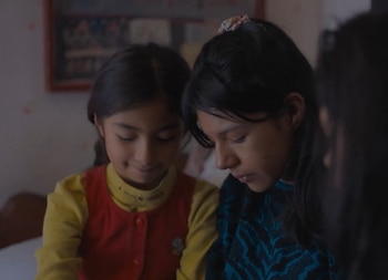 Primer plano de dos niñas jóvenes. La niña de la izquierda, con suéter amarillo y chaleco rojo, sonríe. La de la derecha lleva suéter azul y negro