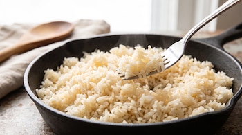 Tenedor de metal levantando arroz blanco recién cocido dentro de una sartén negra de hierro fundido, con una cuchara de madera al fondo.