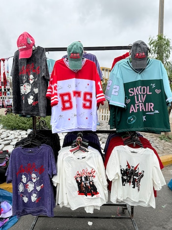 Perchero metálico expuesto en la calle con camisetas, camisetas de béisbol y gorras impresas con fotos y el nombre del grupo BTS