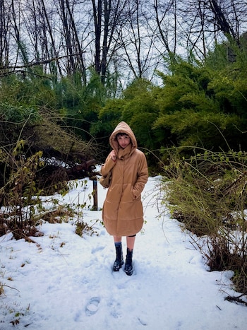 Mujer con campera acolchada beige y botas de cuero negro de pie en un sendero nevado. Rodeada de árboles desnudos y arbustos con hojas verdes