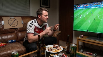 Hombre en camiseta de Alemania mira fútbol en TV. Sentado en sofá con comida, botellas, latas y cartel de 'no hablar'.