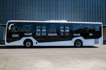 Vista lateral de un moderno autobús eléctrico blanco y negro con el logo "Línea 5 Macro Aeropuerto" y "100% Eléctrico", estacionado frente a una pared metálica
