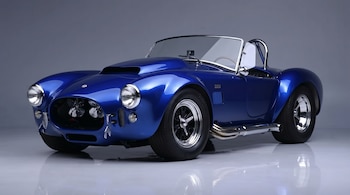 Un automóvil deportivo descapotable azul brillante, modelo Shelby Cobra, se muestra en una vista frontal tres cuartos sobre un fondo gris liso, reflejado en el suelo.