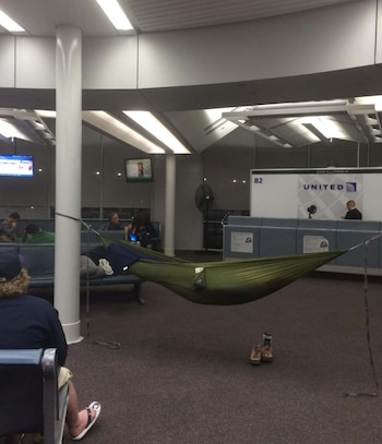 Hombre sentado de espaldas, una hamaca verde colgada de un pilar, personas en bancos, mostrador de United Airlines, pantallas y botas en el suelo de un aeropuerto