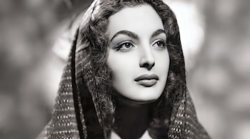 Primer plano de una mujer con cabello oscuro rizado, cejas definidas, ojos grandes y labios maquillados, con fondo difuminado en escala de grises.