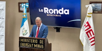 Hombre en traje azul con corbata roja en atril de madera. Pantalla muestra 'La Ronda Ministerial'. Bandera de Guatemala y bandera del Ministerio de Energía y Minas