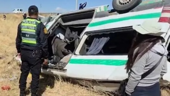 Un agente de la policía de carreteras y una mujer permanecen junto a una minivan volcada de color blanco y verde en un campo seco