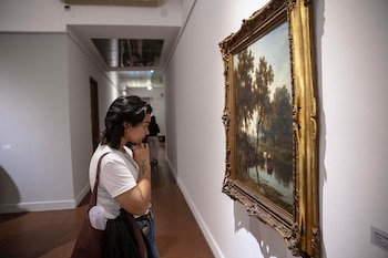 Joven con camiseta blanca y pelo oscuro mira de cerca una pintura enmarcada en oro de un paisaje con árboles y un río en una galería