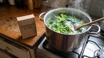 Olla de acero inoxidable con brócoli, guisantes y espinacas hirviendo en una estufa; a su lado, caja de bicarbonato de sodio sobre encimera de madera.