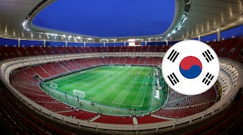 Vista aérea de un estadio de fútbol moderno con asientos rojos y campo verde brillante, iluminado bajo un cielo azul oscuro. Una bandera de Corea del Sur en un círculo está superpuesta en la imagen.