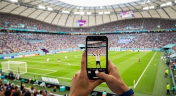 Manos sosteniendo un smartphone en un estadio de fútbol abarrotado. La pantalla del teléfono muestra a un jugador de fútbol celebrando un gol.