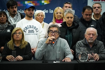 Grupo de personas, mayormente adultos, sentados y de pie detrás de una mesa negra. Un hombre mayor en el centro habla por un micrófono; otros observan con seriedad