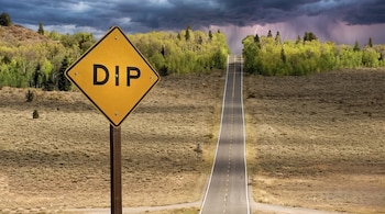 Señal de tráfico amarilla con la palabra "DIP" en un poste, una carretera pavimentada, colinas con árboles y un cielo tormentoso con relámpagos.