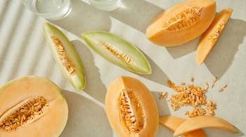 Vista aérea de mitades y rodajas de melones de pulpa naranja y verde con semillas, sobre una superficie clara con sombras y vasos.