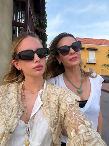 Dos mujeres con gafas de sol, collares y ropa de calle. Detrás se aprecian fachadas de edificios de colores y un cielo claro
