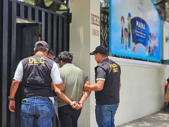 Dos agentes policiales con chalecos negros sujetan a un hombre esposado de camiseta verde frente a una reja negra