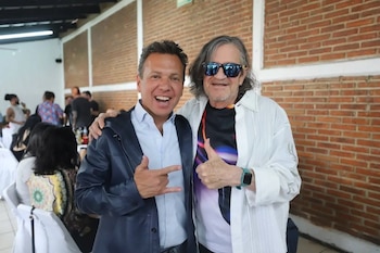 Primer plano de dos hombres sonriendo, uno con chaqueta azul y el otro con camisa blanca y gafas de sol. Hacen gestos con las manos. Fondo de pared de ladrillo