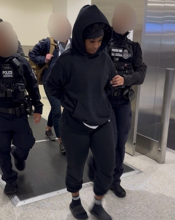 Una mujer con sudadera negra y pantalón deportivo es escoltada por dos agentes uniformados en un interior. Otras personas transitan