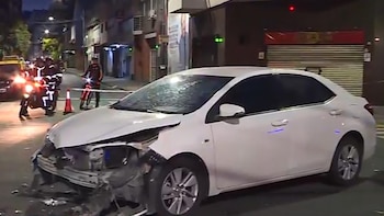 Vista nocturna de un sedán blanco con el paragolpes y el capó delantero destrozados en una calle urbana oscura. Se aprecian luces de vehículos y agentes de seguridad a lo lejos