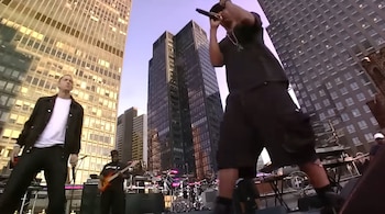 Eminem, en camisa blanca y chaqueta negra, en un escenario exterior con rascacielos de fondo. Otro artista canta en primer plano, con un bajista visible
