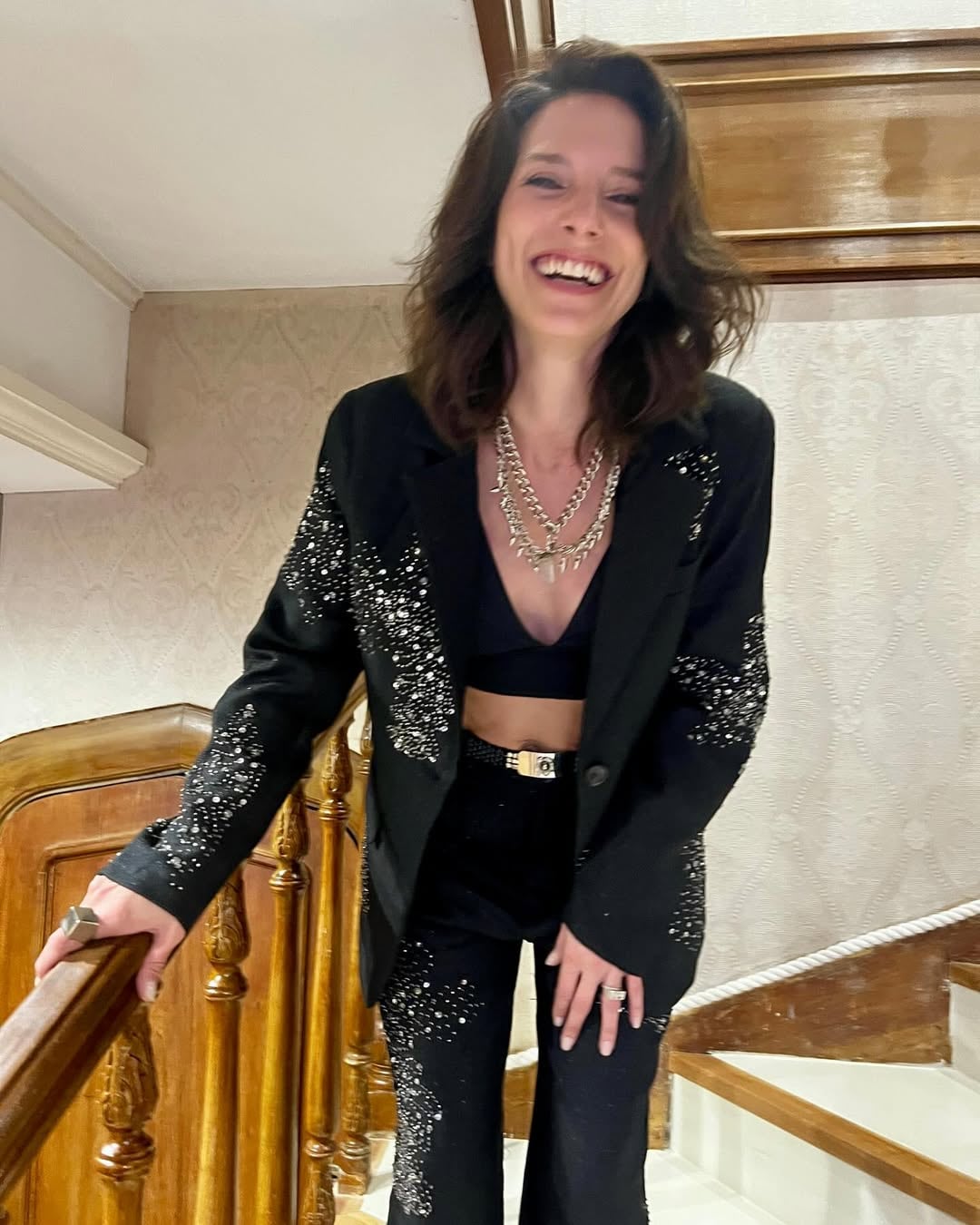 Manuela Pal sonríe con alegría luciendo un elegante conjunto negro con detalles brillantes mientras posa en una escalera de madera.