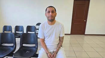 Hombre con camiseta y pantalón blancos, esposas, tatuajes en brazo, sentado en silla negra en sala con paredes blancas, piso de baldosas y puerta de madera.
