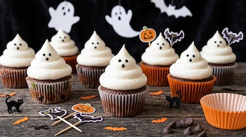 Muffins con cobertura blanca decorada como fantasmas sobre una mesa de madera junto a figuras de papel de murciélagos, gatos y calabazas.
