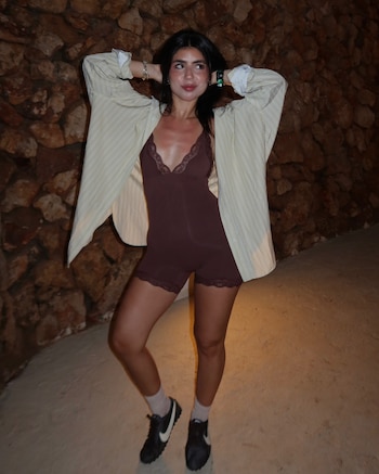 Mujer joven de cabello oscuro con romper marrón, camisa a rayas, calcetines claros y zapatillas deportivas, de pie junto a una pared de piedra