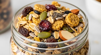 Granola variada en frasco de vidrio con avena, almendras, arándanos, pasas, y semillas de girasol, calabaza y chía sobre fondo blanco.