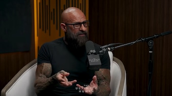 Un hombre calvo con barba, gafas y tatuajes gesticula frente a un micrófono en un estudio con paneles de madera