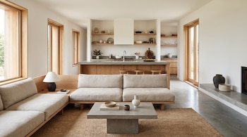 Vista frontal de una sala de estar minimalista con sofás claros, mesa de centro de hormigón y alfombra de yute, conectada a una cocina con estantes de madera y ventanas.