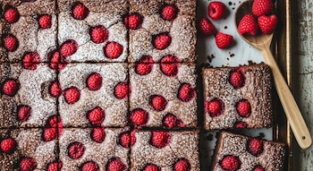 Vista cenital de brownies de chocolate con frambuesas y azúcar glas cortados en cuadrados, en una bandeja, con una cuchara de madera y frambuesas sueltas.
