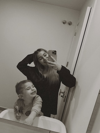 Reflejo en blanco y negro de una mujer sonriente con teléfono y un niño riendo que sostiene un cepillo de dientes. Se ve un lavabo y una puerta