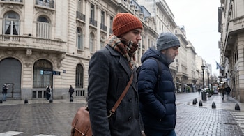 Vista frontal de dos hombres jóvenes con gorros y abrigos gruesos caminando por una calle adoquinada bordada por edificios antiguos en un día gris.