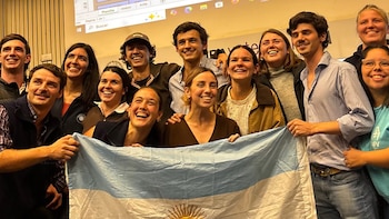 Un grupo de aproximadamente quince jóvenes, sonrientes y riendo, sostiene una bandera argentina en primer plano. Hay un hombre con bigote en la izquierda y una pantalla detrás