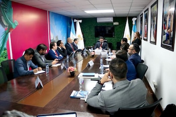 Una sala de reuniones con una larga mesa de madera y unas quince personas sentadas. Un hombre con barba habla por micrófono. Hay banderas de Guatemala y un quetzal en la pared