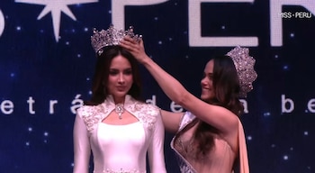 Dos mujeres en un escenario. Una, con vestido blanco y tiara, baja la mirada mientras otra, sonriendo, le coloca una corona brillante. Fondo oscuro con estrellas