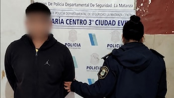 Un hombre con el rostro difuminado permanece de pie junto a una oficial de policía frente a una pared con carteles institucionales