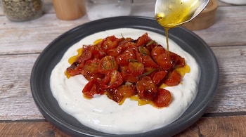 Un plato gris contiene una capa de crema blanca y tomates cherry asados con hierbas. Una cuchara vierte aceite de oliva encima. Fondo de madera.
