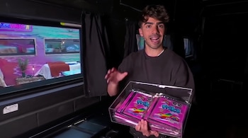 Hombre sonriente con barba y camiseta oscura sostiene bandeja con paquetes rosados de "Friks Arcoíris Pop" frente a una pantalla con escena de habitación y cartel "VIBES".