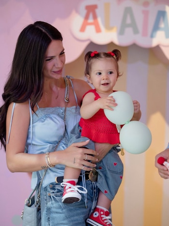Una mujer de cabello oscuro sostiene a una niña con zapatillas rojas que sostiene dos globos pequeños ante un cartel con el nombre Alaia