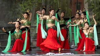 Grupo de siete mujeres asiáticas en el escenario, vistiendo tops verdes bordados, faldas rojas amplias y tocados dorados, con brazos en poses de danza