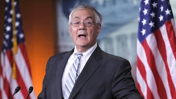 Un hombre blanco mayor con gafas y cabello gris, Barney Frank, vistiendo un traje y corbata rayada, habla con la boca abierta frente a dos banderas estadounidenses