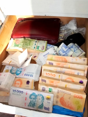 Primer plano de un cajón con grandes fajos de billetes de pesos argentinos de diversas denominaciones, una cartera roja y bolsas transparentes