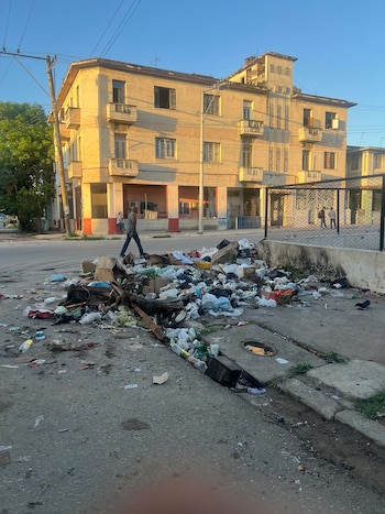 Vista de una calle de La Habana con una gran pila de basura en primer plano. Un hombre camina a la izquierda, y un edificio antiguo de color claro se ve al fondo