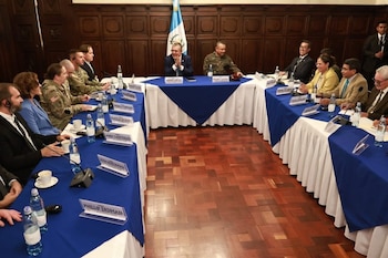 Vista amplia de una reunión formal en una sala con paredes de madera y bandera guatemalteca. Personas en trajes y uniformes sentadas alrededor de una mesa en forma de U