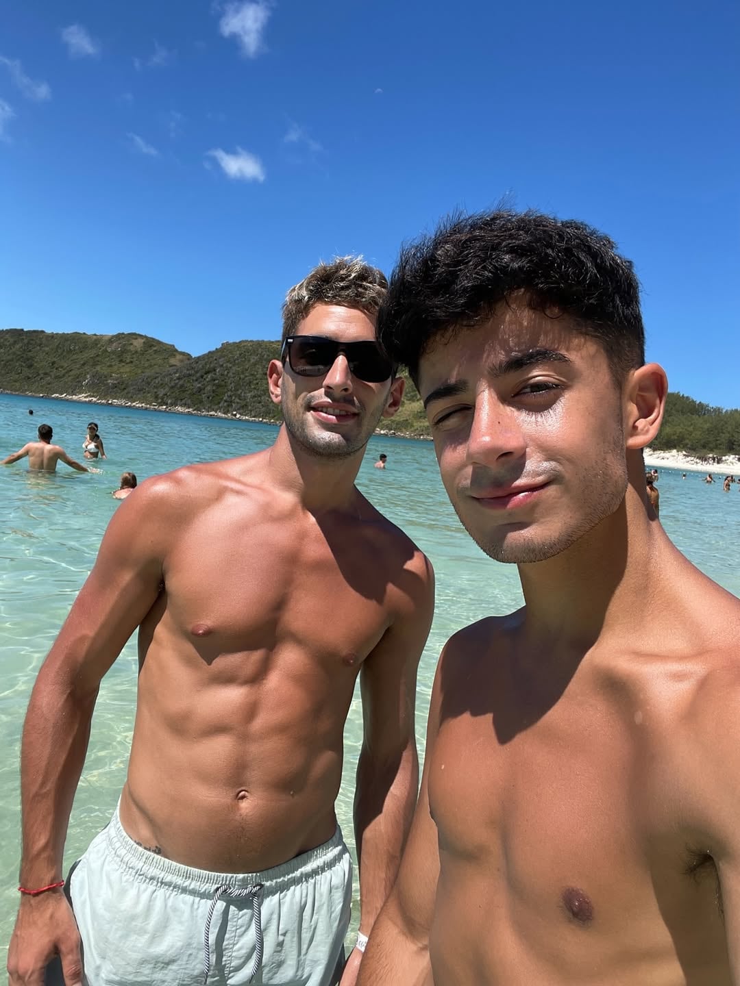 Kevsho e Iván en una playa paradisíaca de aguas cristalinas bajo un sol radiante, tomando un merecido descanso